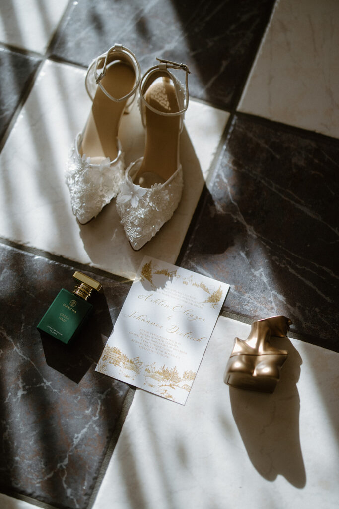 asheville wedding photographer A pair of white lace bridal shoes, a green perfume bottle, a gold abstract sculpture, and a wedding invitation card arranged on a checkered tile floor with soft sunlight, captured beautifully by an Asheville wedding photographer.