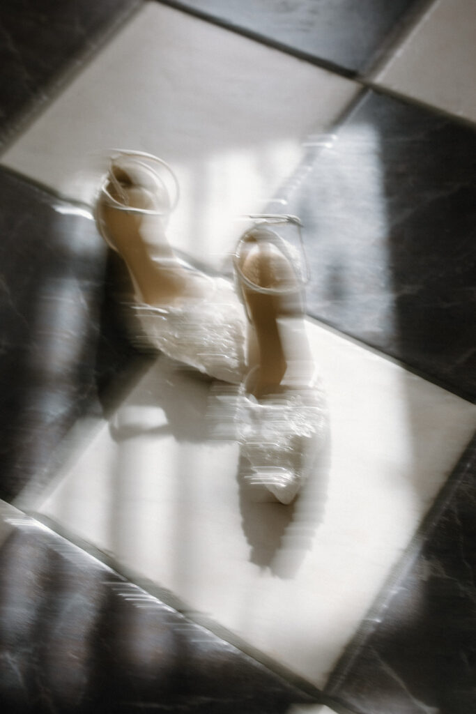 asheville wedding photographer A pair of elegant high-heeled shoes with ankle straps and embellishments sits on a black-and-white checkered floor, bathed in soft, diffused light—captured with the dreamy artistry of an Asheville wedding photographer.