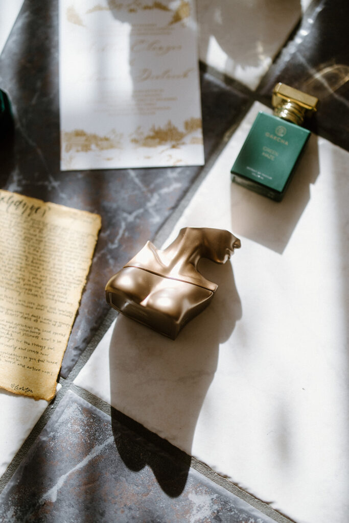 asheville wedding photographer A gold sculpture of a torso, a green perfume bottle, an old handwritten letter, and a decorative white paper lie on sunlit tiles, casting distinct shadows—captured with the artistry of an Asheville wedding photographer.