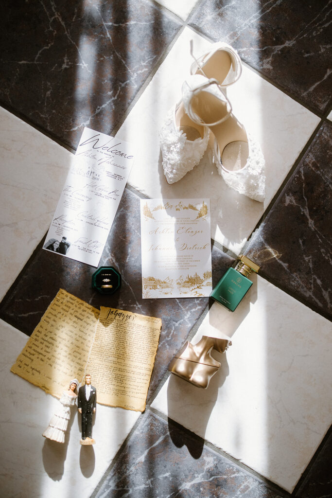 asheville wedding photographer Flat lay of wedding items on tiled floor, captured by an Asheville wedding photographer—lace bridal shoes, invitations, green perfume bottle, gold rings in a box, old letter, and bride and groom figurine. Light shines diagonally.