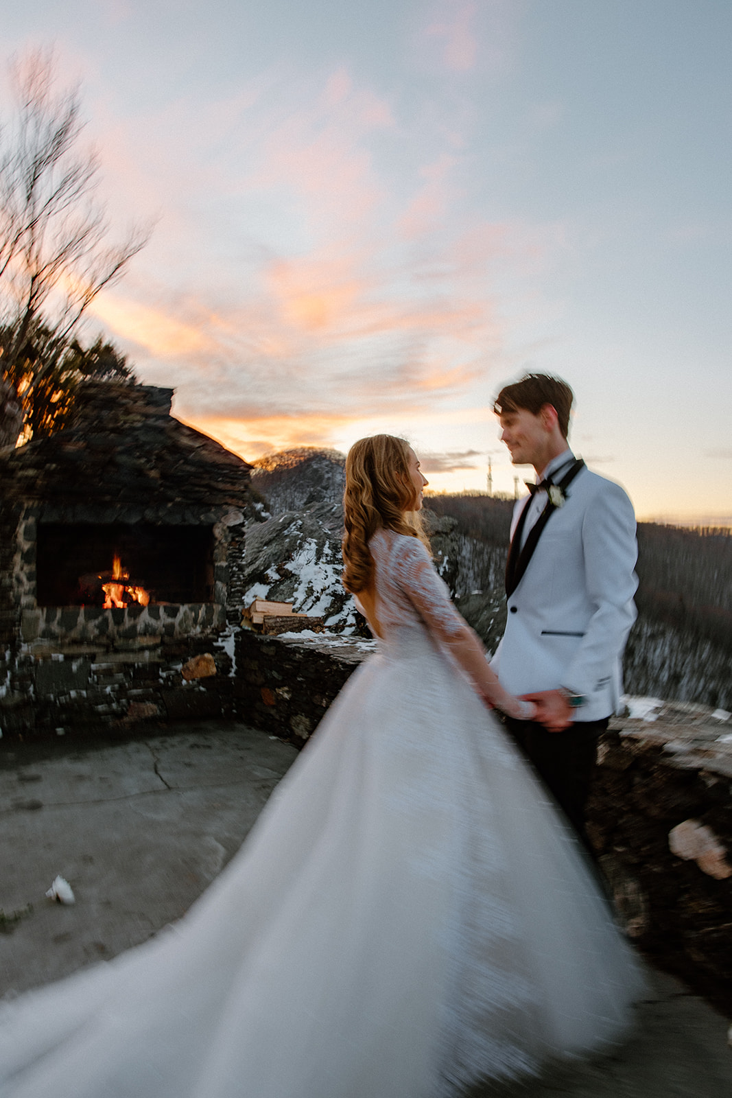 asheville wedding photographer A bride in a long white gown and a groom in a white tuxedo jacket stand outdoors holding hands at sunset, with a stone fireplace and snowy mountains behind them—captured perfectly by an Asheville wedding photographer.