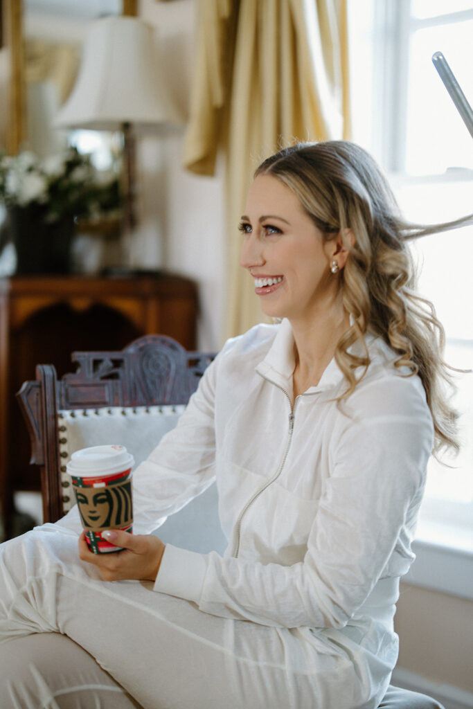 asheville wedding photographer A woman in white loungewear sits smiling on a chair, holding a coffee cup with a festive sleeve. Captured by an Asheville wedding photographer, she’s in a warmly lit room with a window, lamp, and flowers in the background.