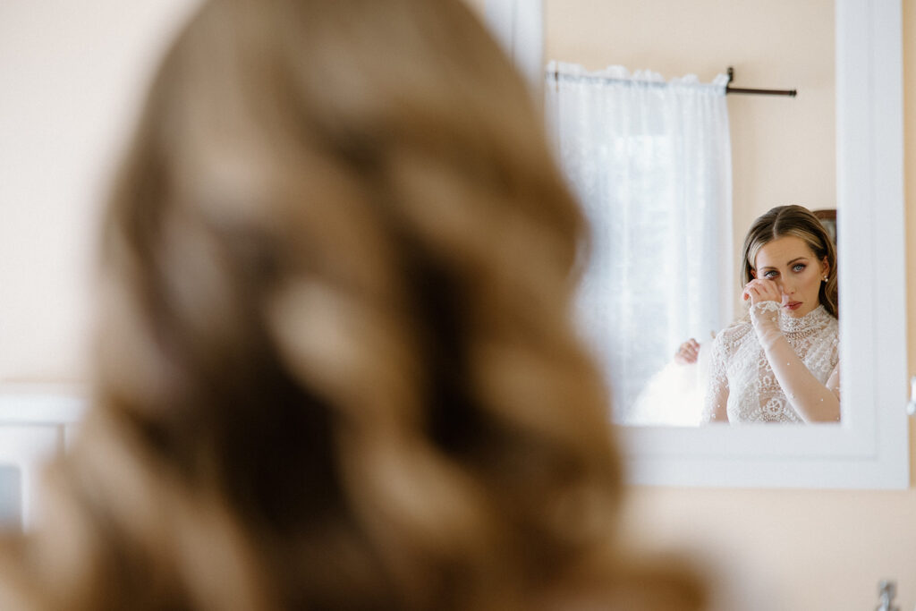 asheville wedding photographer A woman in a lace dress is reflected in a mirror, holding her hand near her face as if wiping tears, while the back of her wavy brown hair is visible in the foreground—a moment beautifully captured by an Asheville wedding photographer.
