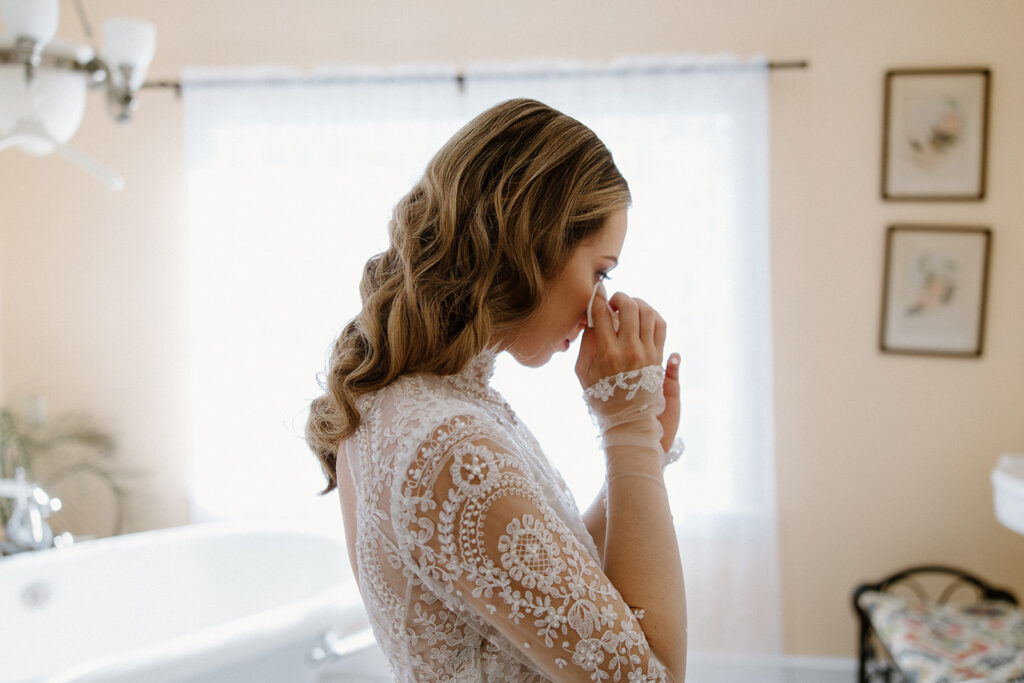 asheville wedding photographer A bride in a lace wedding dress stands in a softly lit room, wiping a tear from her eye, with light streaming through a window behind her—captured beautifully by an Asheville wedding photographer.