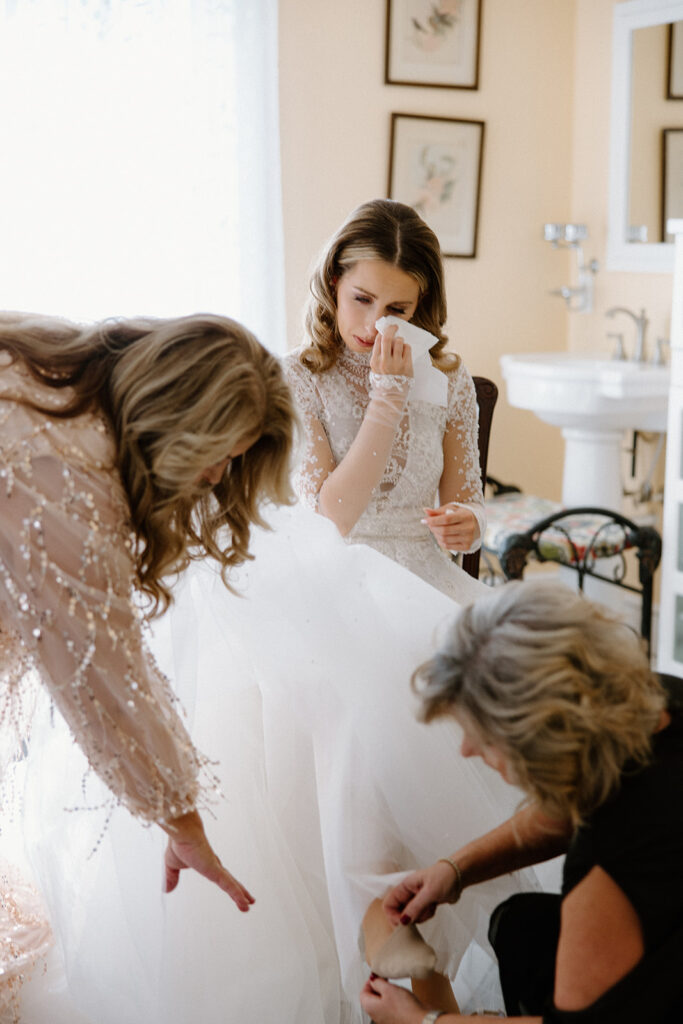 asheville wedding photographer A bride in a white gown wipes a tear from her eye as two women help adjust her dress. Captured by an Asheville wedding photographer, the softly lit room features framed art on the walls and a sink in the background.