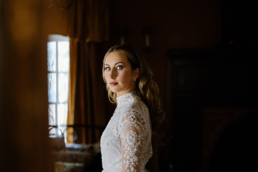 asheville wedding photographer A woman in a white, lace, long-sleeved dress stands indoors by a window with golden curtains, looking over her shoulder toward the camera in soft, natural light—captured by an Asheville wedding photographer.