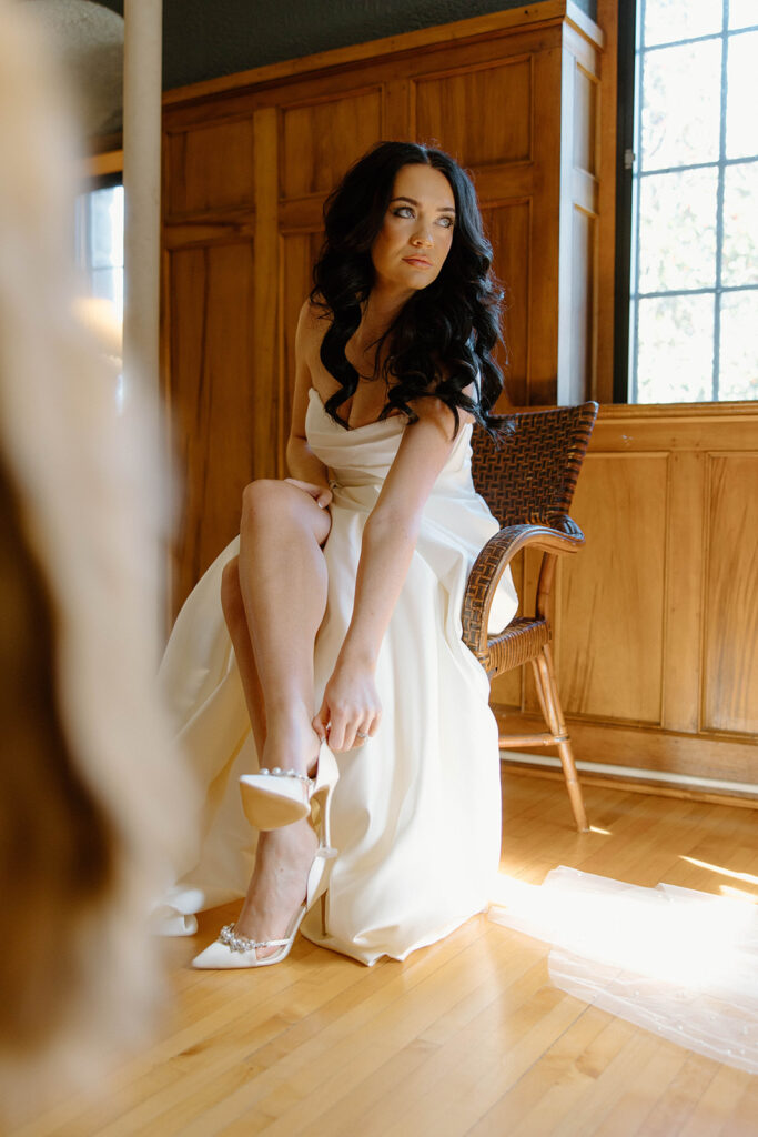 asheville wedding photographer A woman in a white dress sits on a wooden chair, adjusting her white high-heeled shoe. Sunlight streams in from a nearby window, illuminating the wooden floor—perfect inspiration for any Asheville wedding photographer.
