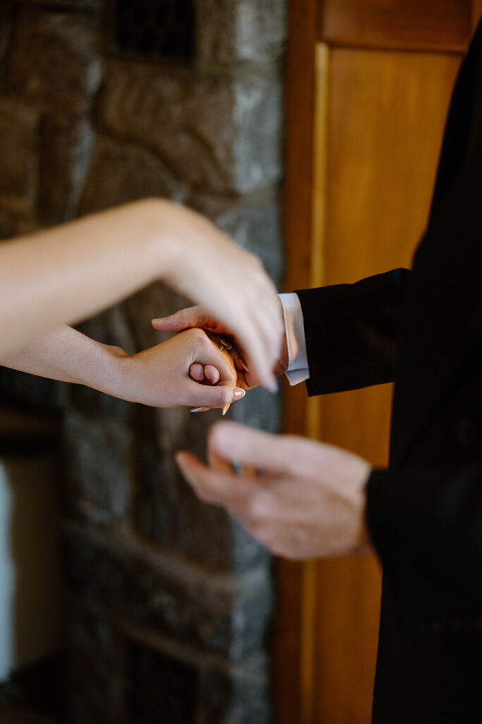 asheville wedding photographer Two people holding hands, one in a black suit and the other in light attire, stand indoors near a stone wall and wooden door. Captured by an Asheville wedding photographer, only their hands and arms are visible, suggesting a formal or intimate moment.