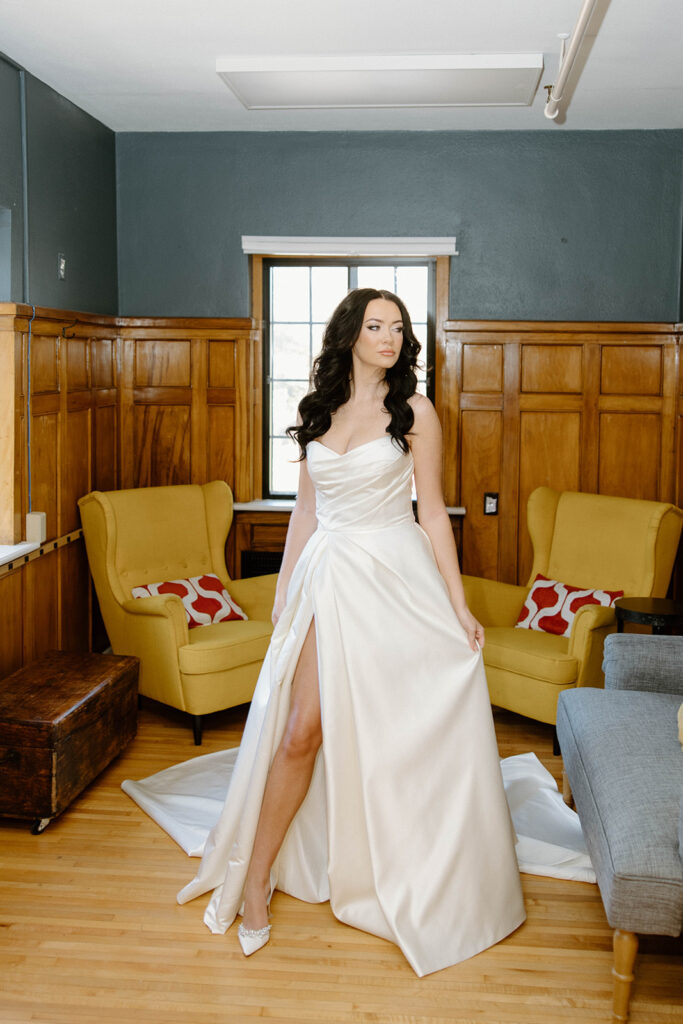 asheville wedding photographer A woman in a white wedding dress with a thigh-high slit stands in a room with wood paneling, yellow armchairs, and natural light streaming through the window—captured beautifully by an Asheville wedding photographer.