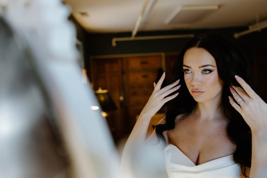 asheville wedding photographer A woman with long dark hair and an off-the-shoulder white dress looks at herself in the mirror, adjusting her hair and showing a ring on her finger in a softly lit room, captured beautifully by an Asheville wedding photographer.