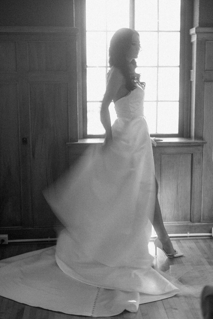 asheville wedding photographer A bride in a flowing white gown stands by a sunlit window, her dress swirling as soft light streams in, creating a dreamy effect in this black and white photograph by an Asheville wedding photographer.