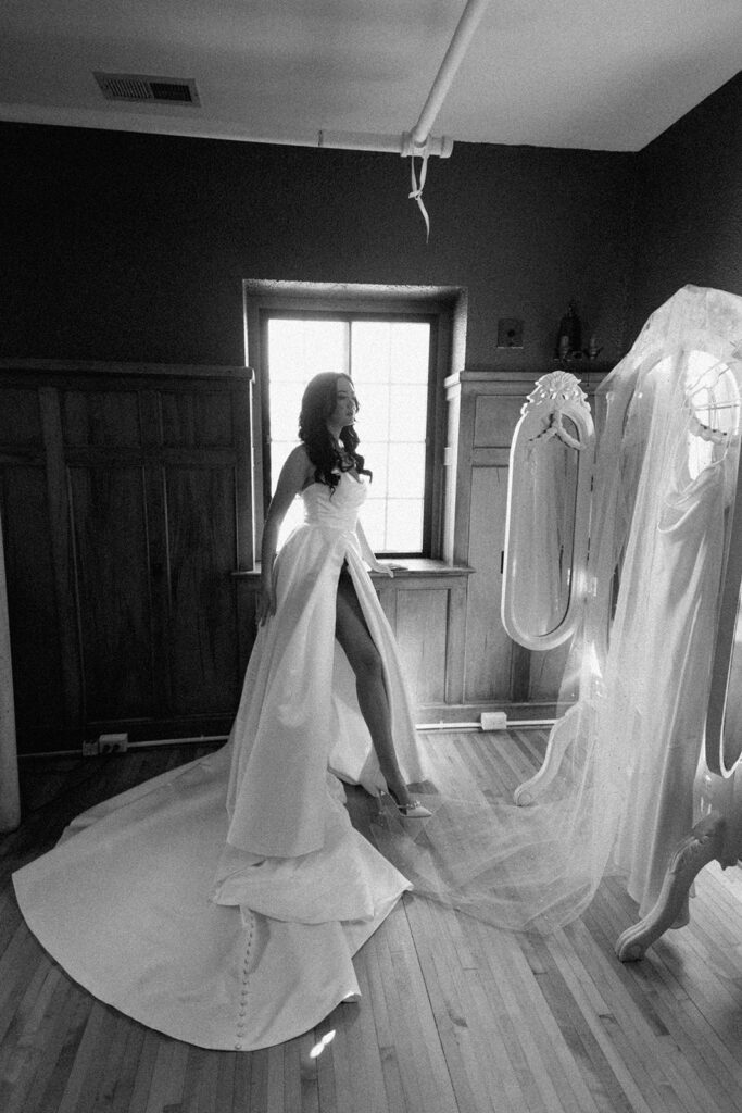 asheville wedding photographer A woman in a strapless wedding dress poses beside a window in a wooden-paneled room, her long train flowing behind her. Captured by an Asheville wedding photographer, the black and white image features her veil draped over a standing mirror.