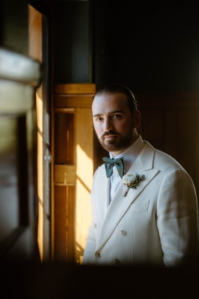 asheville wedding photographer A man with a beard and slicked-back hair, wearing a white suit with a green bow tie and boutonniere, stands by a window in a warmly lit wooden room, looking into the camera—captured by an Asheville wedding photographer.