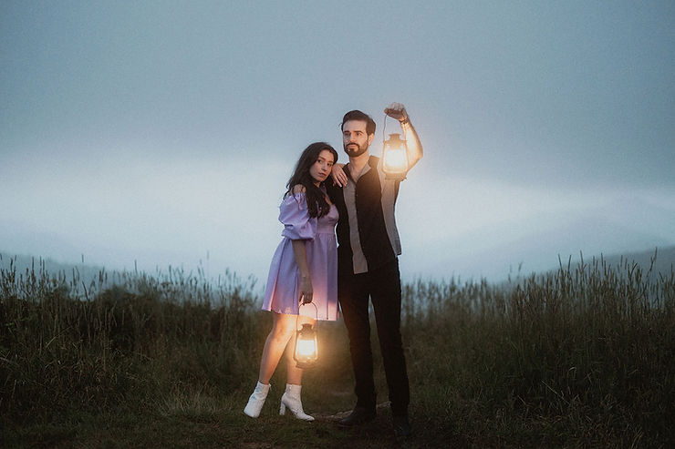 asheville wedding photographer A woman in a purple dress and a man in dark clothes stand in a grassy field at dusk, each holding a lit lantern, as an Asheville wedding photographer captures the misty hills and cloudy sky in the background.