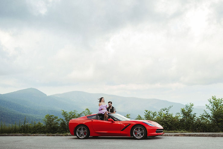 asheville wedding photographer Two people sit on the roof of a red sports car parked beside a road, with green mountains and cloudy skies in the background—an adventurous moment perfectly captured by an Asheville wedding photographer.