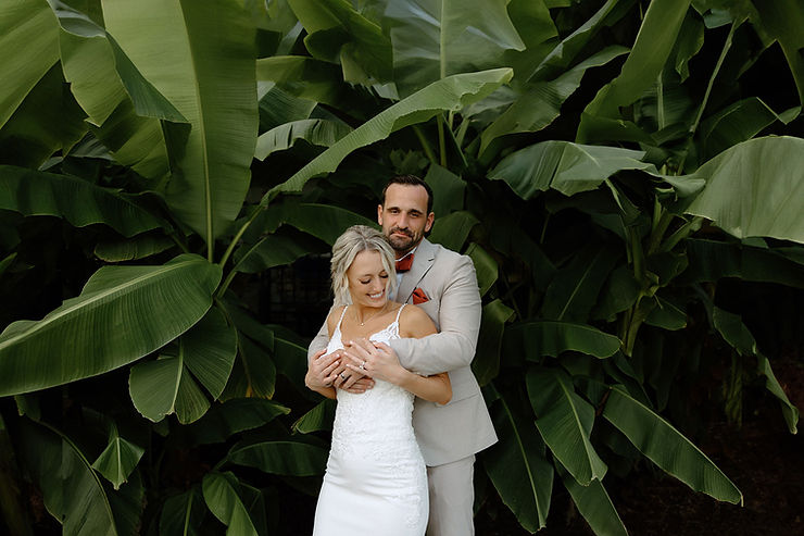 asheville wedding photographer A bride in a white dress and a groom in a light suit embrace and smile while standing in front of large green tropical leaves, captured beautifully by an Asheville wedding photographer.