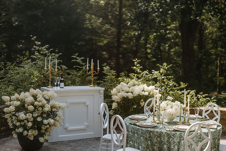 asheville wedding photographer An elegant outdoor table setting with white chairs, a green and white patterned tablecloth, white floral arrangements, and tall candlesticks—perfect inspiration for an Asheville wedding photographer. A white bar with candlesticks and greenery sits nearby amid lush trees.