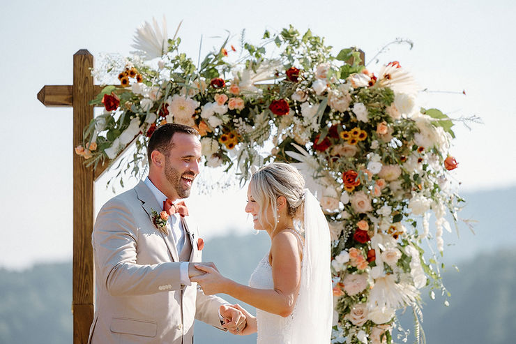 asheville wedding photographer A bride and groom smile and hold hands in front of a floral arch during an outdoor wedding ceremony on a sunny day, beautifully captured by an Asheville wedding photographer.
