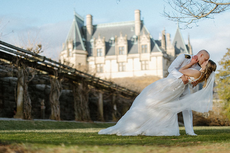 asheville wedding photographer A groom in a white military uniform dips and kisses his bride in a flowing white gown on a grassy lawn, with a grand castle-like building in the background, beautifully captured by an Asheville wedding photographer.