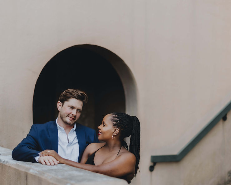 asheville wedding photographer A couple stands on a concrete staircase, gazing at each other and smiling. Captured by an Asheville wedding photographer, the man wears a blue suit while the woman in a black dress holds his arm. A round archway and beige wall complete the scene.
