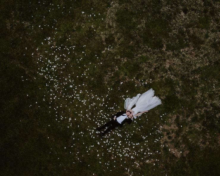 asheville wedding photographer A bride and groom lie on grass, surrounded by scattered white flower petals. Captured from above by an Asheville wedding photographer, the bride’s long veil and white dress contrast beautifully with the groom’s dark suit.