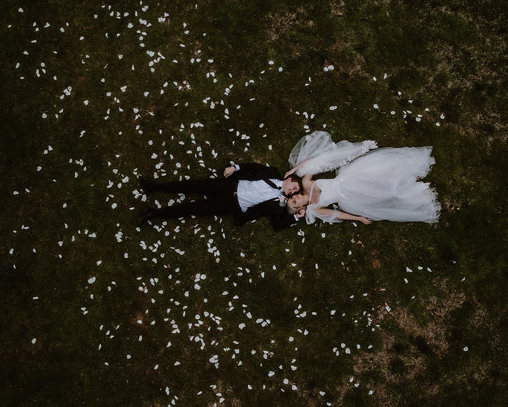 asheville wedding photographer A bride and groom in wedding attire lie side by side on grass, surrounded by scattered white flower petals, viewed from above by an Asheville wedding photographer.