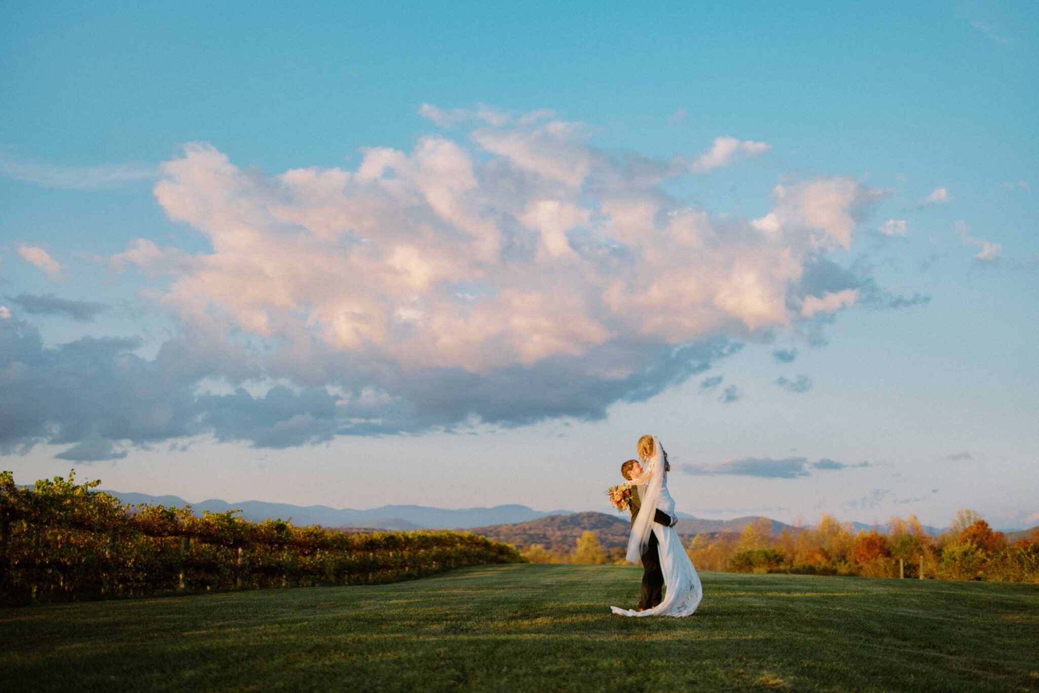 asheville wedding photographer A bride and groom embrace on a grassy field at sunset, with dramatic clouds and distant mountains—an Asheville wedding photographer captures this romantic and serene outdoor scene.