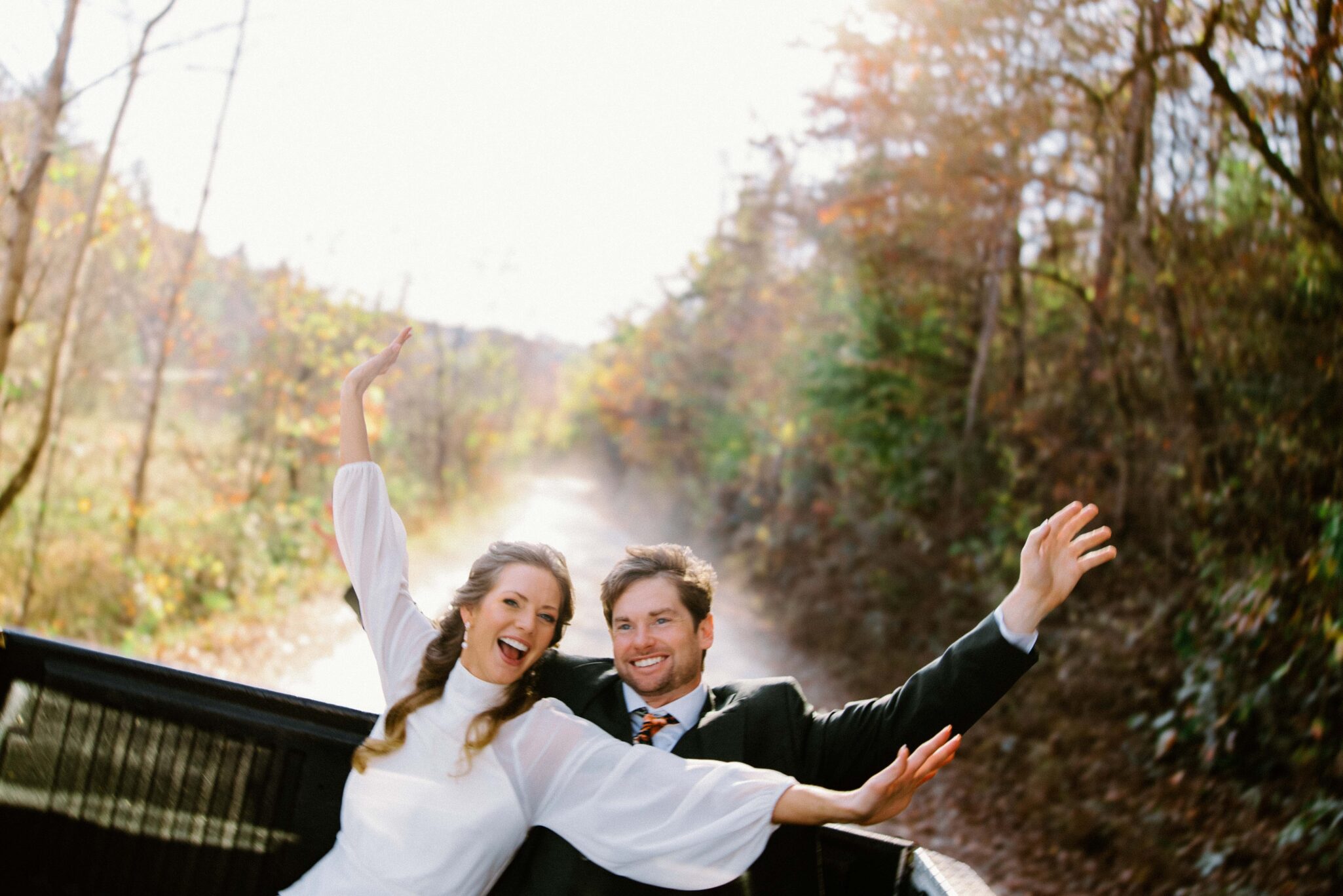asheville wedding photographer A joyful bride and groom in wedding attire sit in the back of a truck, smiling and raising their arms as they ride down a sunlit, tree-lined country road—captured perfectly by an Asheville wedding photographer.