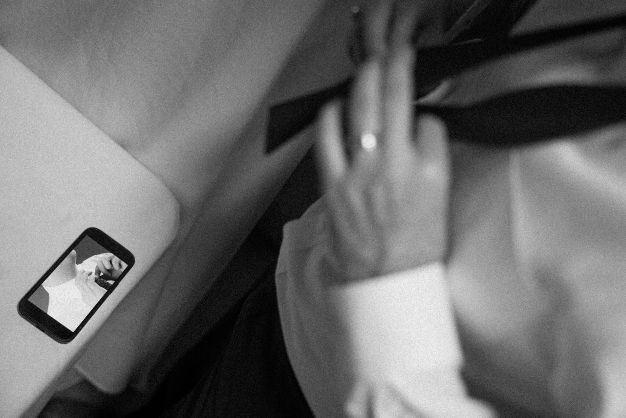 asheville wedding photographer A person in a dress shirt ties a necktie, their reflection captured on a smartphone screen nearby. This black-and-white image evokes the classic style often favored by an Asheville wedding photographer.