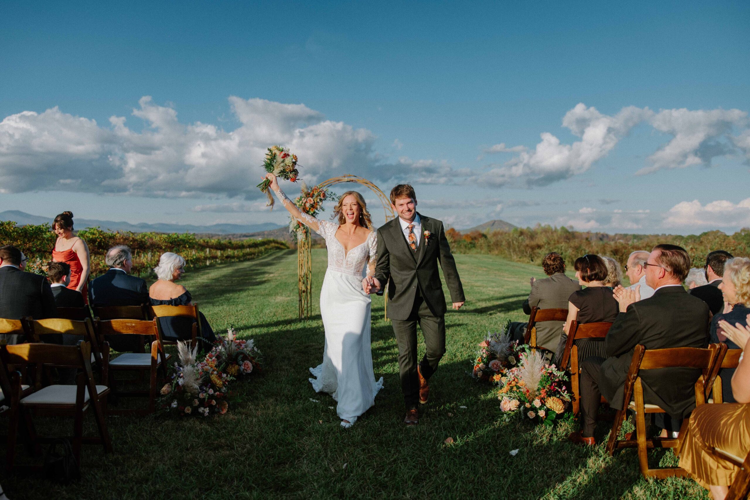 asheville wedding photographer A bride and groom walk joyfully down the aisle outdoors after their wedding ceremony, holding hands as guests smile and clap. Captured by an Asheville wedding photographer, the bride raises her bouquet in celebration against a scenic, sunny landscape.