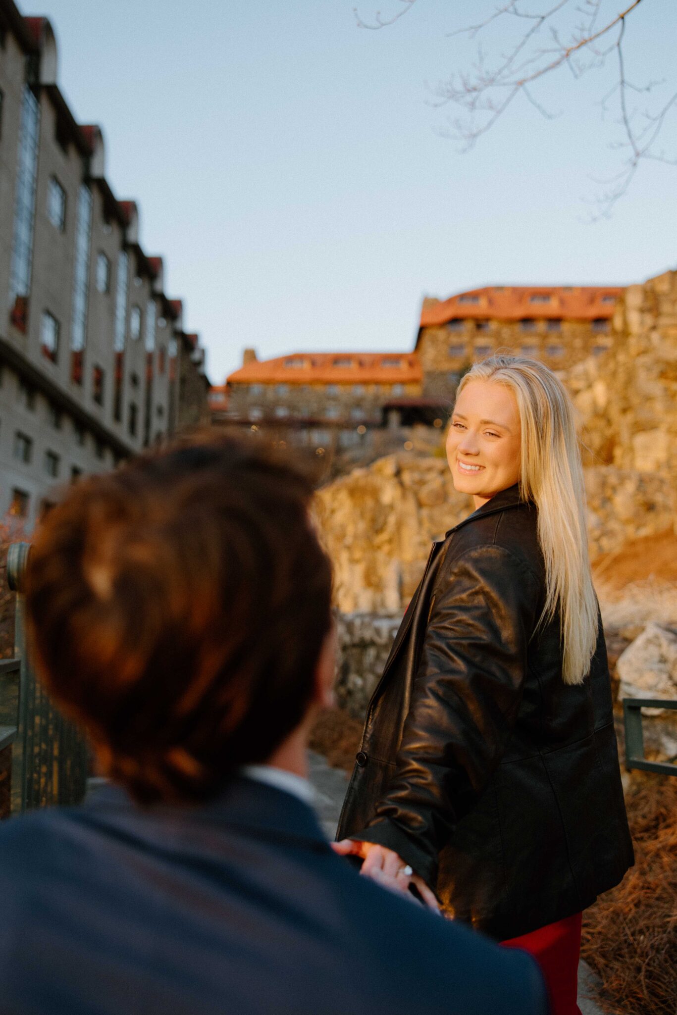 asheville wedding photographer A woman with long blonde hair in a black jacket smiles back while holding hands with a person in a suit, walking outdoors near stone buildings at sunset—captured beautifully by an Asheville wedding photographer.