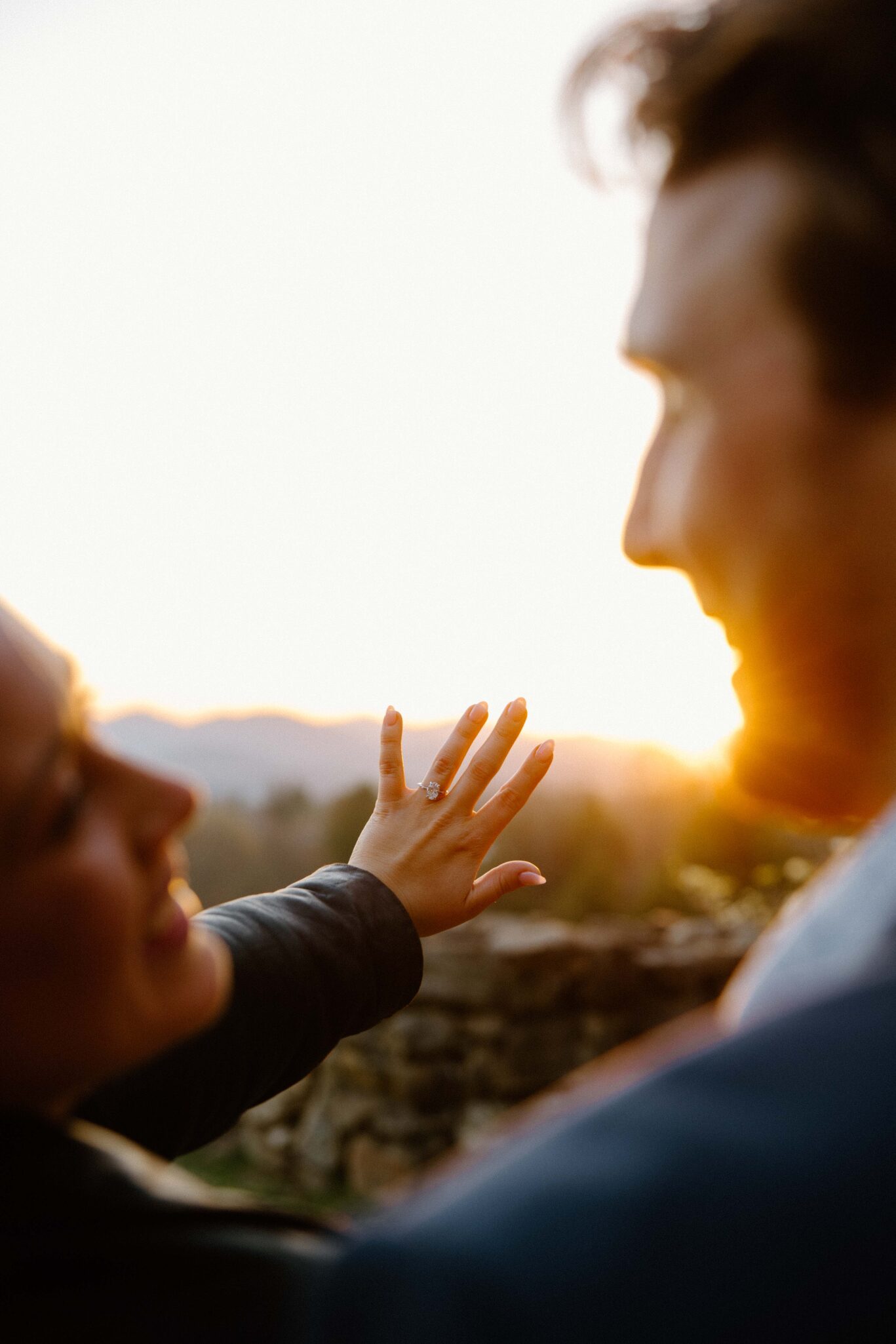 asheville wedding photographer A person holds up their hand to show an engagement ring while smiling at another. Captured by an Asheville wedding photographer, the couple stands outdoors with a sunset and mountain backdrop, bathed in warm, soft light.