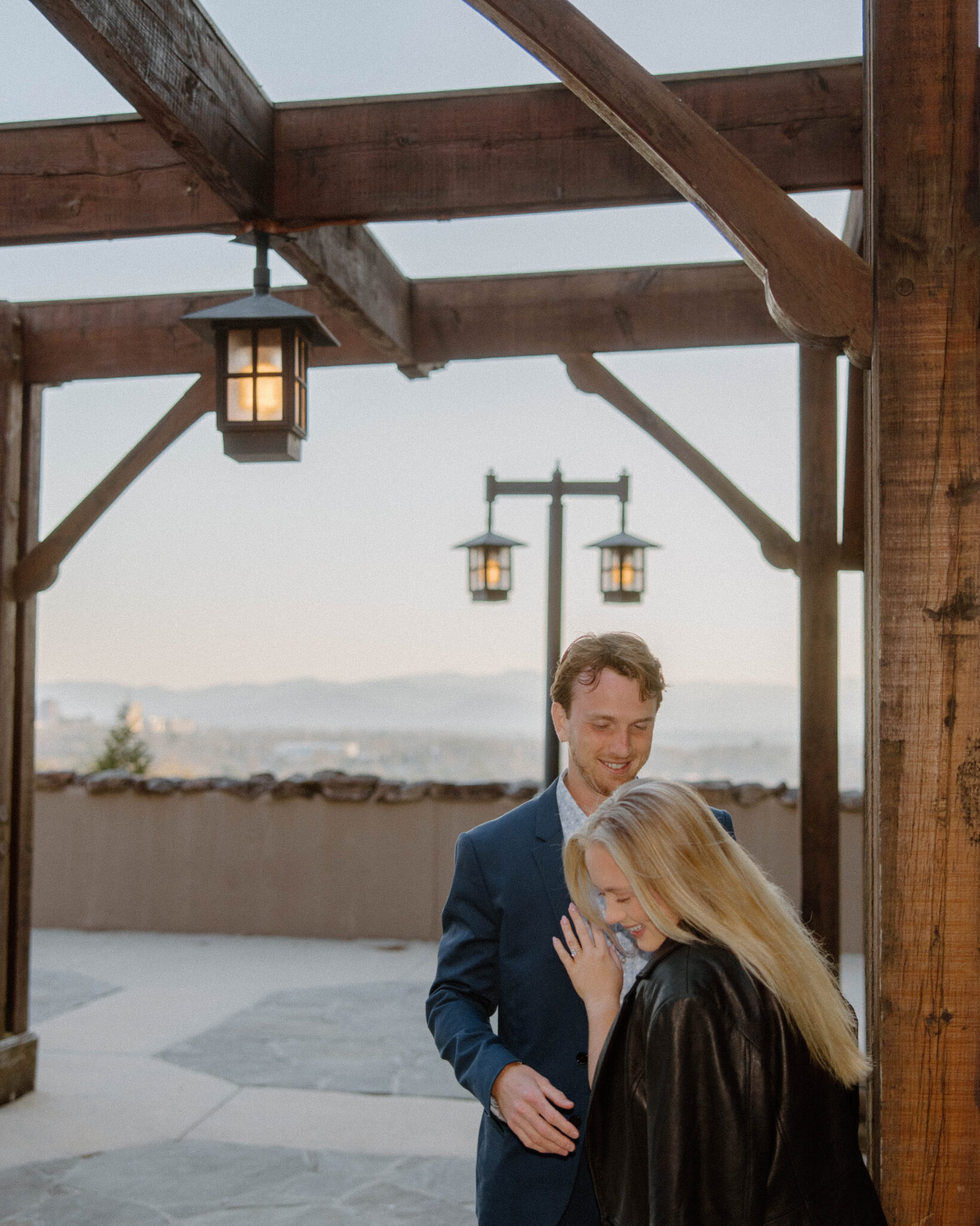 asheville wedding photographer A man in a suit and a woman in a black jacket smile and laugh together under a wooden pergola with hanging lanterns, with mountains and a stone path behind them—captured beautifully by an Asheville wedding photographer.