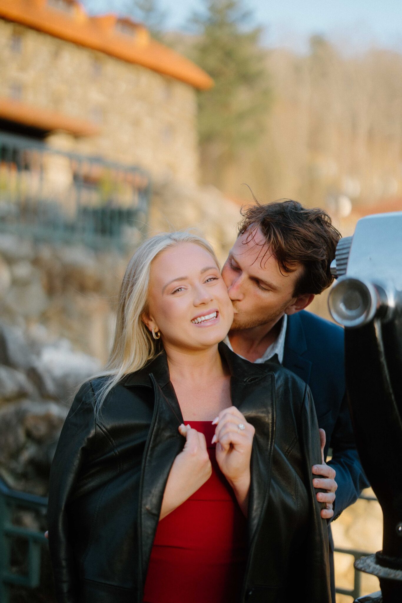 asheville wedding photographer A man gently kisses a smiling woman on the cheek as she holds her jacket. They stand outdoors near stone buildings and a scenic overlook with a coin-operated viewing machine—captured by an Asheville wedding photographer.
