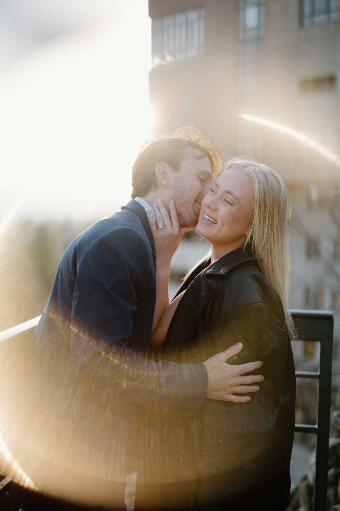 asheville wedding photographer A man in a suit kisses a smiling woman with long blonde hair on the cheek. Captured by an Asheville wedding photographer, they embrace outdoors in soft sunlight, with a circular light flare and a blurred building in the background.