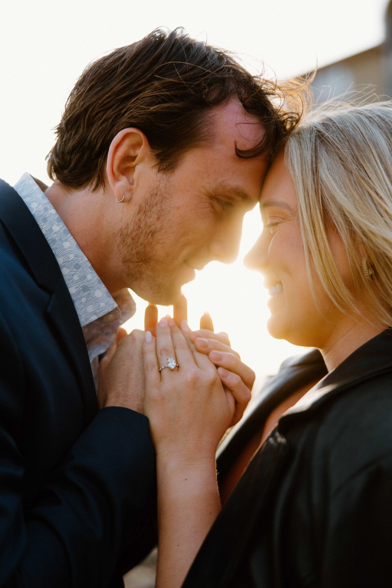 asheville wedding photographer A couple stands close together, smiling and touching foreheads, with hands clasped and the sun shining between them. Captured by an Asheville wedding photographer, the woman proudly displays a ring on her finger.