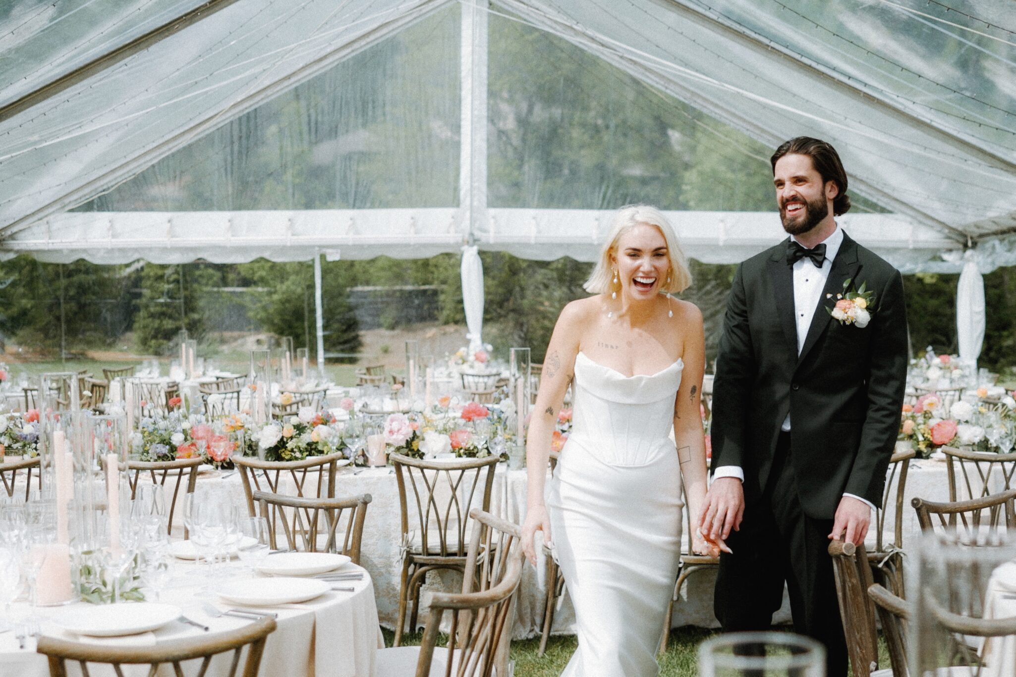 asheville wedding photographer A bride in a white dress and a groom in a black tuxedo smile and hold hands while walking under a clear tent decorated for a wedding reception, surrounded by flowers and greenery—captured perfectly by an Asheville wedding photographer.