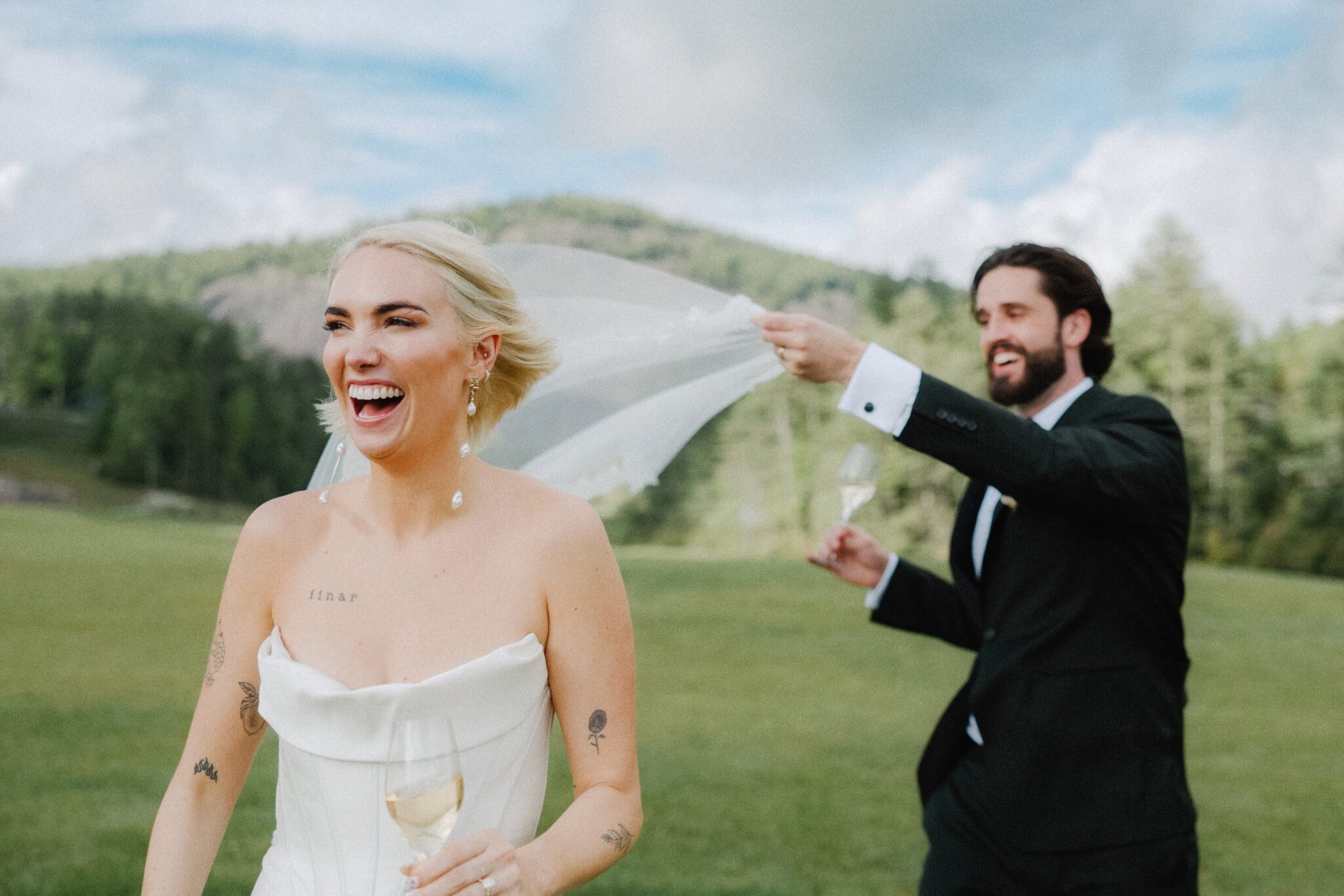 asheville wedding photographer A bride in a strapless white dress laughs while a groom in a black suit playfully holds her veil outdoors, with green hills and trees behind them. Captured by an Asheville wedding photographer, both appear joyful and relaxed.