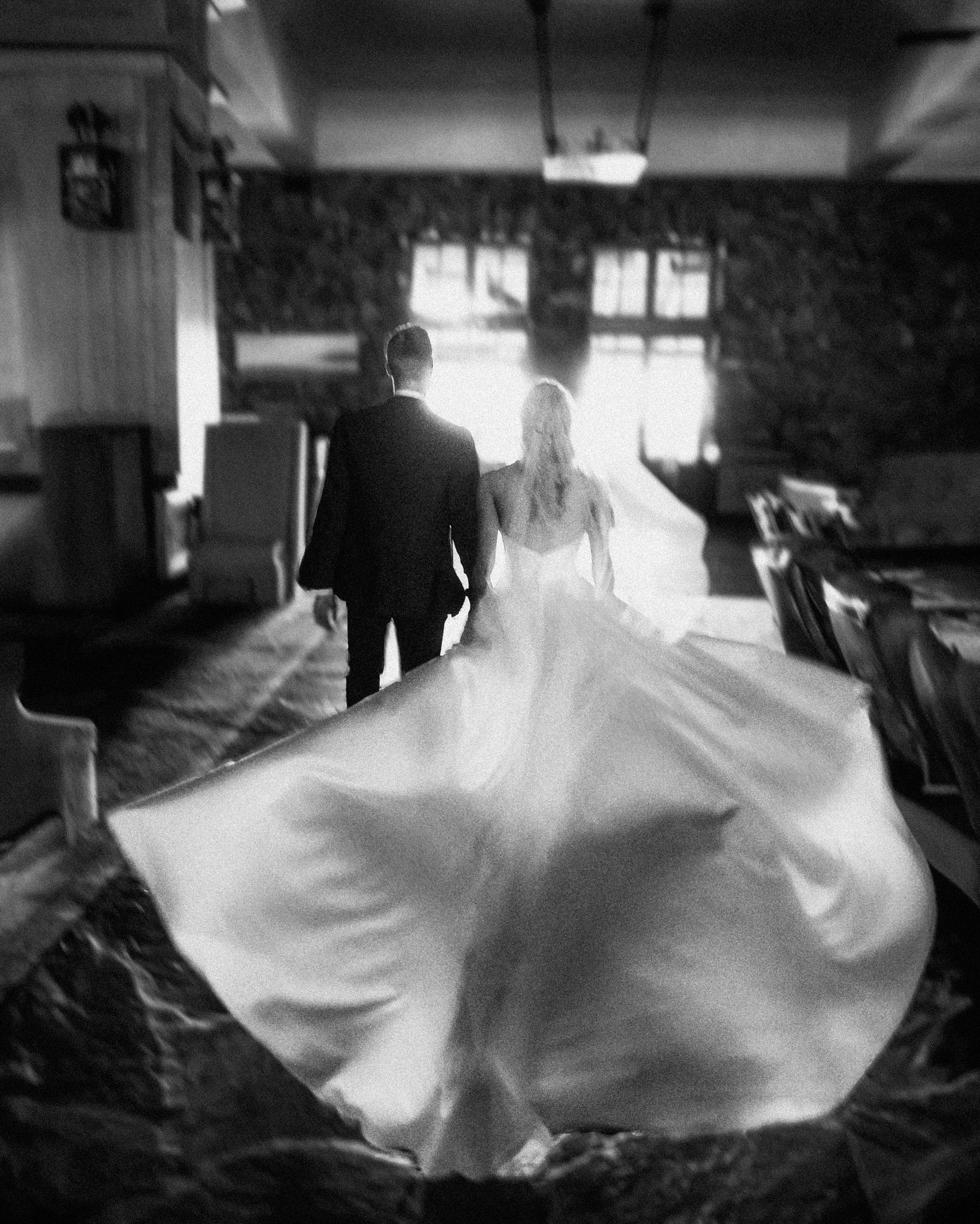 asheville wedding photographer A bride and groom walk hand in hand indoors, with the bride’s flowing dress captured in motion. This black and white image by an Asheville wedding photographer creates a dramatic, romantic atmosphere as light shines through windows in the background.