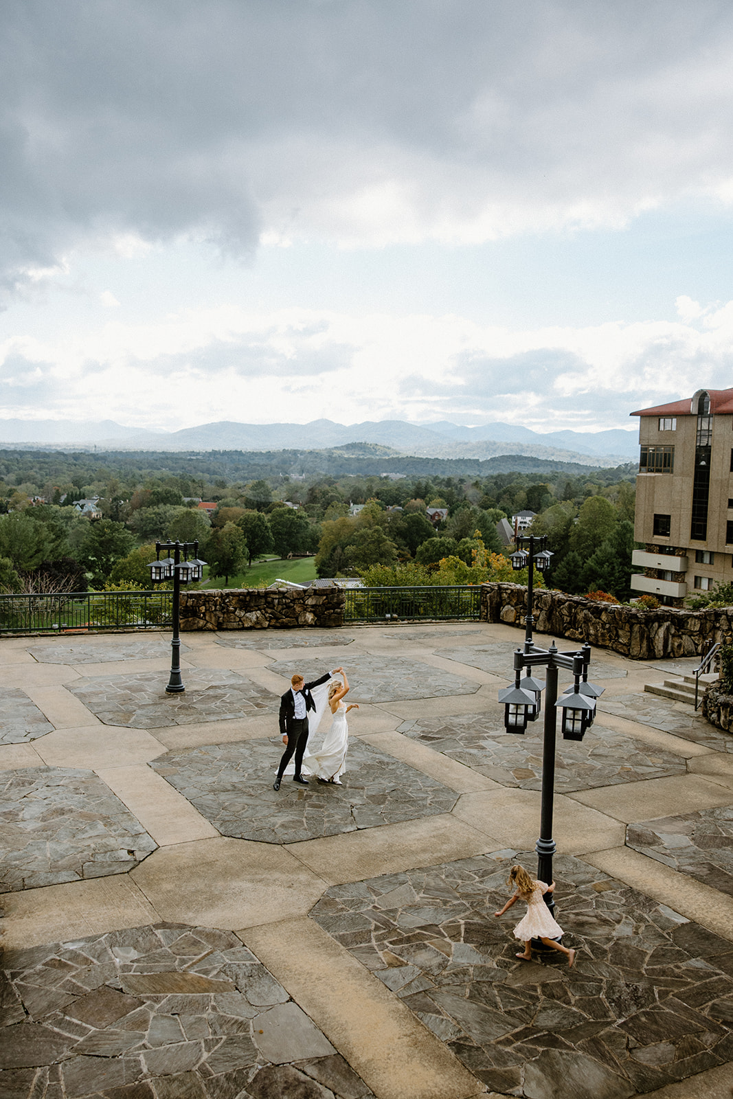 Bride and groom dancing on a wide stone terrace as dark storm clouds gather overhead, with distant Blue Ridge Mountains in the background and a flower girl running nearby.