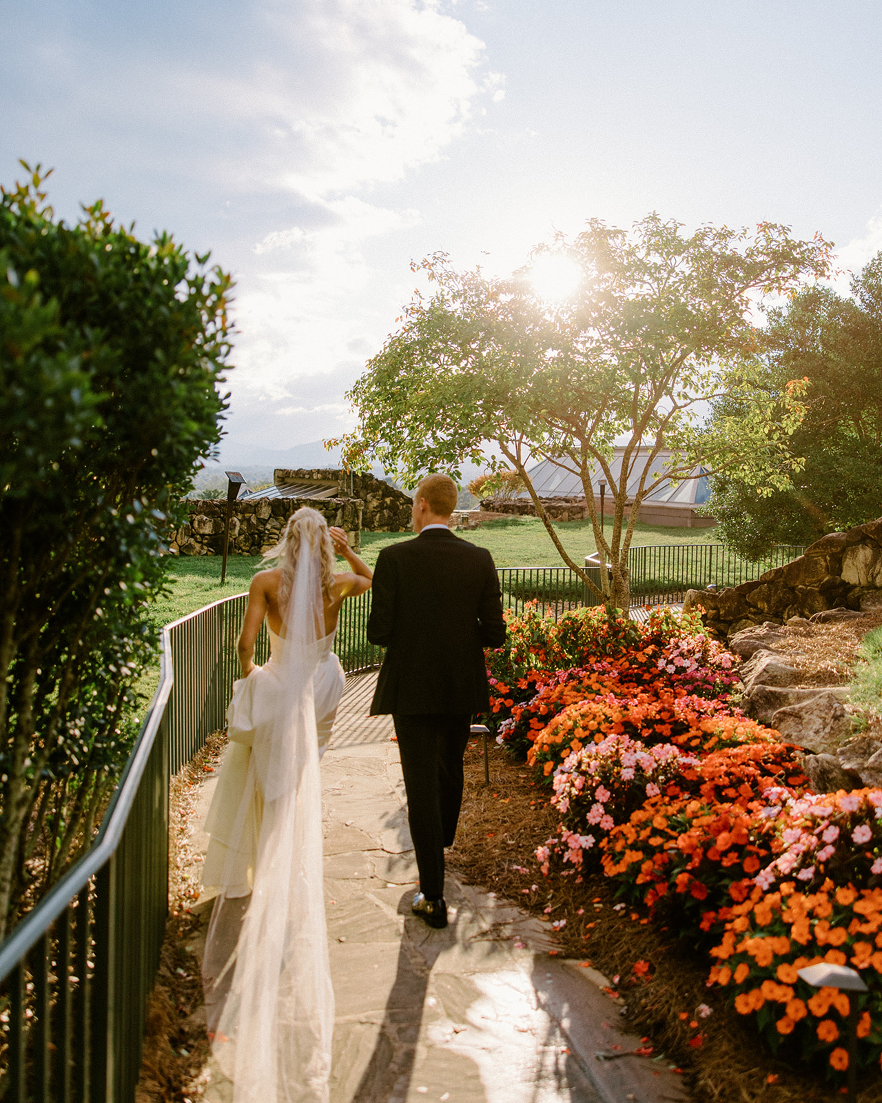 Bride and Groom walk through a sunlit garden near the spa at The Omni Grove Park inn with lots of color as bride adjusts her veil. 