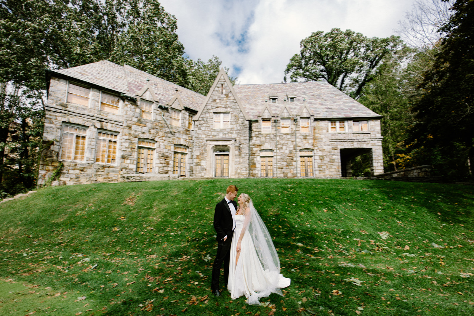 asheville wedding photographer A bride and groom stand closely together on a grassy lawn in front of a stone mansion with tall windows and a pointed roof. Captured by an Asheville wedding photographer, the surrounding trees and sky frame this perfect wedding day moment.