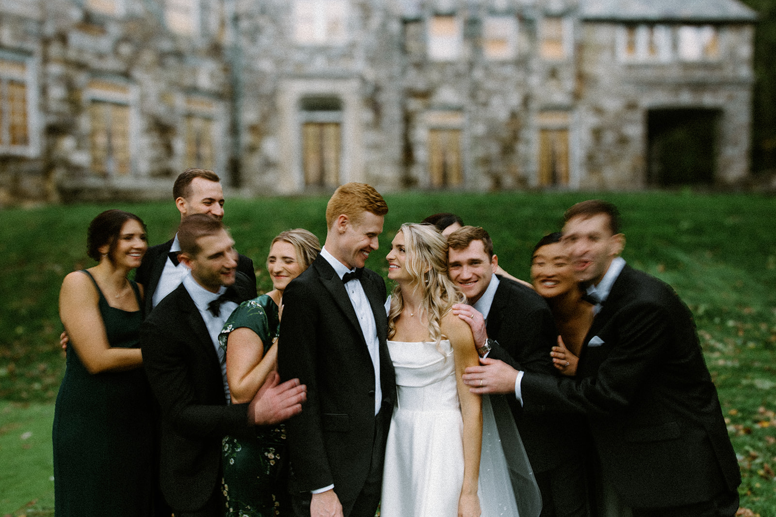 asheville wedding photographer A bride and groom stand smiling outside a stone building, surrounded by a joyful wedding party in dark suits and green dresses, all laughing together—captured perfectly by an Asheville wedding photographer.