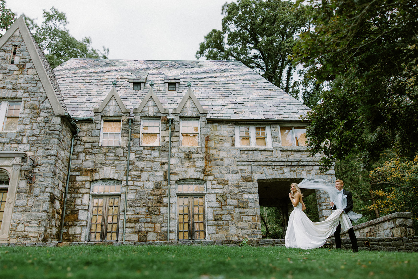asheville wedding photographer A bride and groom stand outside a large stone mansion as their veil flows in the wind. Captured by an Asheville wedding photographer, the moment is set amid lush green grass and trees surrounding the elegant, historic building.