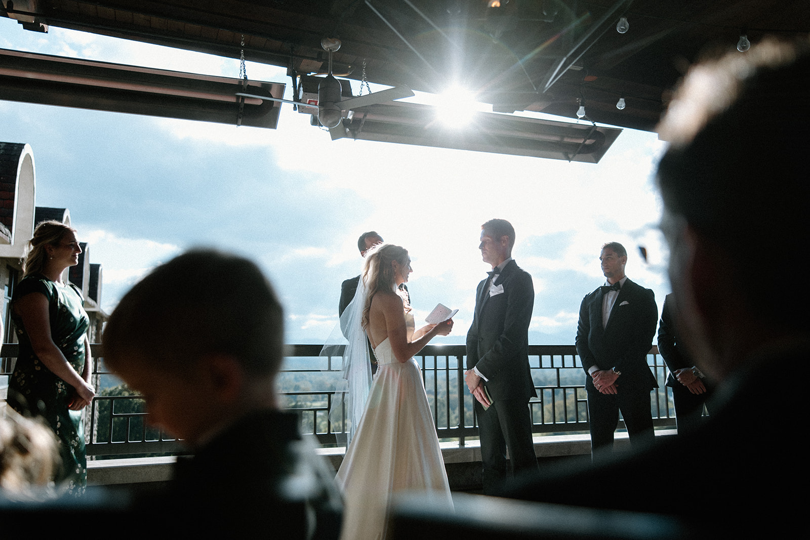 asheville wedding photographer A bride and groom stand facing each other during an outdoor ceremony, with sunlight streaming through the open roof. Bridesmaids, groomsmen, and guests look on as an Asheville wedding photographer captures the magical moment.