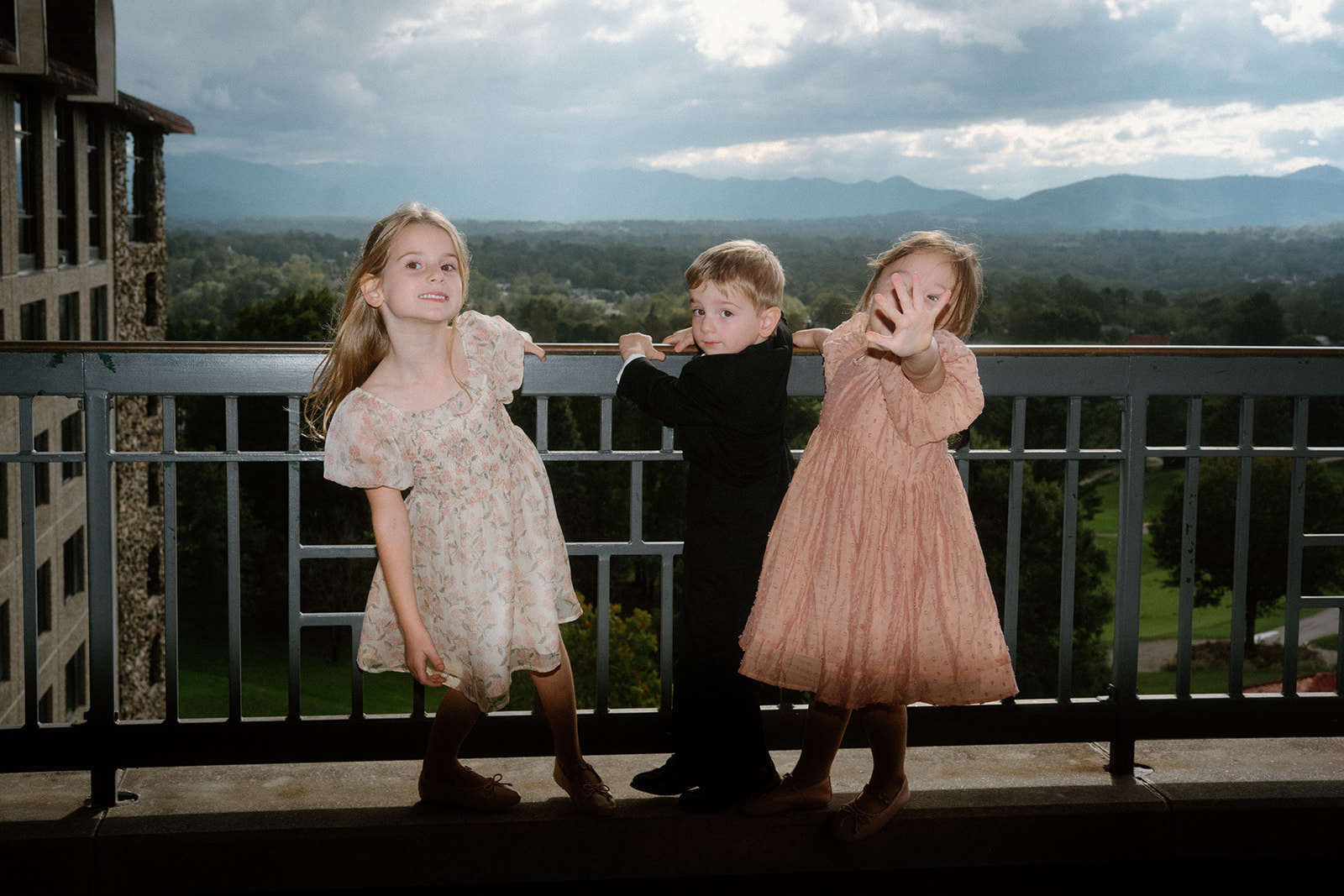 Three flower girls and ring bearer playfully pose on a balcony at The Omni Grove Park Inn, with the Blue Ridge Mountains and dramatic post-hurricane skies in the background.