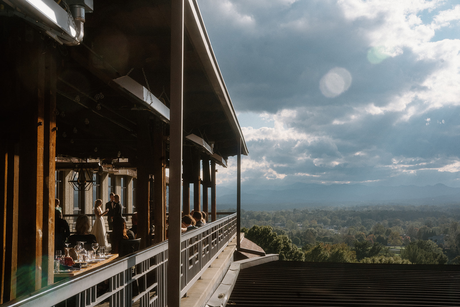 asheville wedding photographer People sit at tables on a covered outdoor balcony overlooking a scenic valley with distant mountains under a partly cloudy sky; sunlight creates lens flares, perfect for an Asheville wedding photographer to capture the moment.
