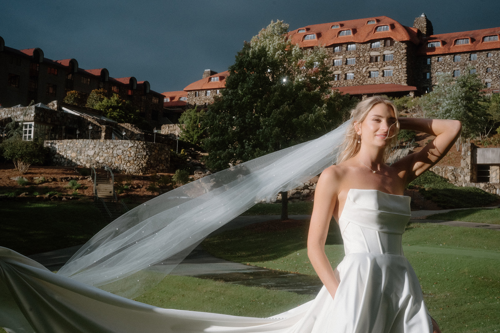 asheville wedding photographer A bride in a strapless white gown poses outdoors, holding her flowing veil, as an Asheville wedding photographer captures the moment against a large stone building and lush green lawn under dramatic sunlight.