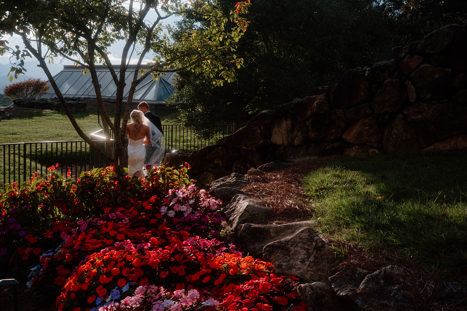 Bride and groom walk hand-in-hand through vibrant flower gardens outside The Grove Park Inn in Asheville, North Carolina, sunlight breaking through after Hurricane Helene passed.