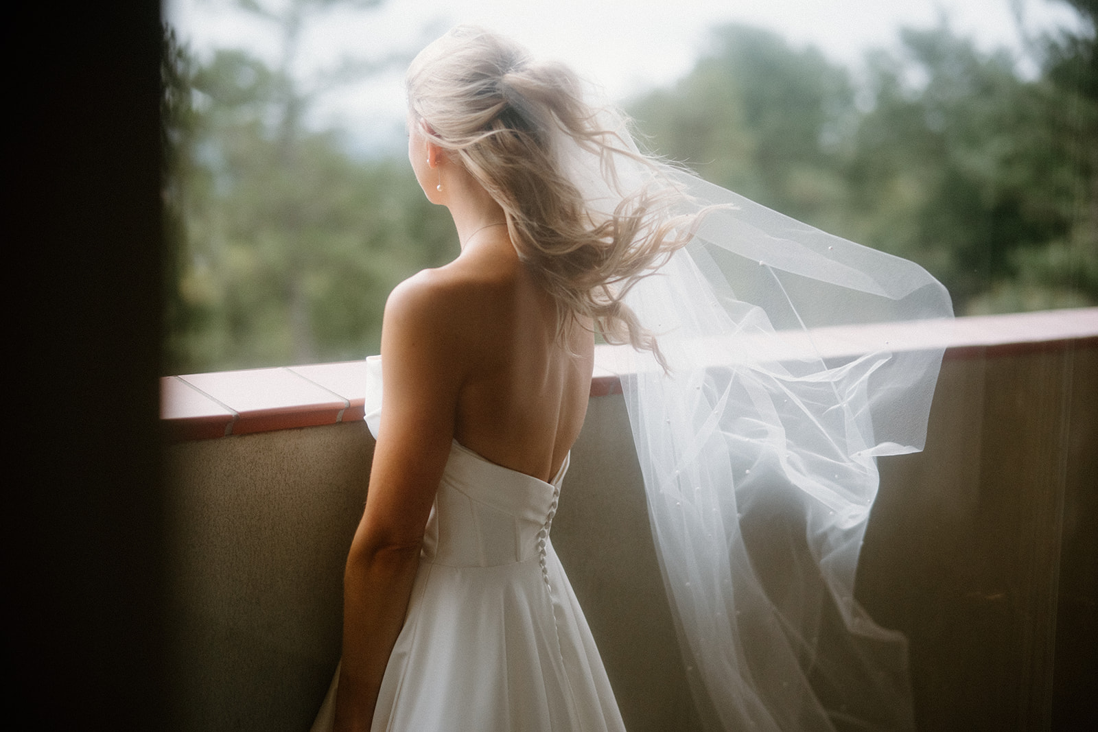 asheville wedding photographer A bride with long blond hair and a flowing veil stands on a balcony, looking out at a blurred, green outdoor landscape. Captured by an Asheville wedding photographer, she wears a strapless white wedding dress.